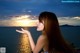 A woman with long red hair standing by the water at sunset.